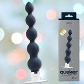 A black silicone-textured VeDO 'Quaker' Anal Vibrator and Prostate Massager with multiple bulb shapes and a white base is displayed alongside its packaging. The packaging prominently showcases the product name and features a gold emblem. The background has a soft bokeh effect with pastel colors, enhancing the appearance of the device's vibration modes.